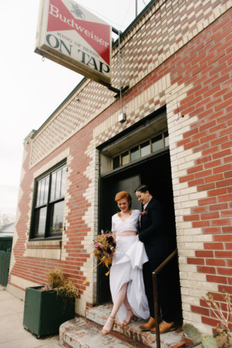 queer couple coming out of a bar in wedding attire. one woman in a suit, one woman in a two piece dress. the woman in a dress is holding a bouquet made of dried flowers.